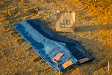 Фото светло-синих, темно-синих джинсов и бумажного пакета с логотипом Woodcutter на фоне желтого грунта карьера