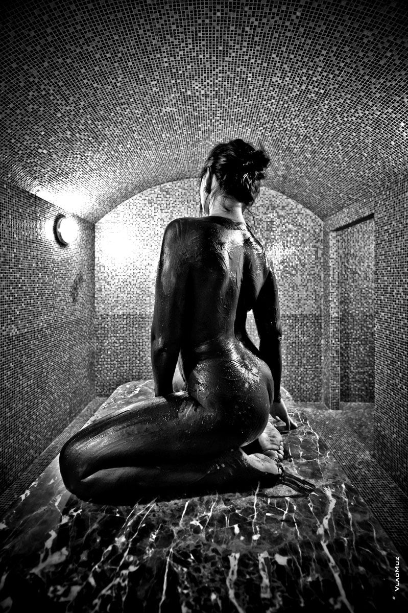 Черно-белое фото обнаженной девушки, обмазанной черной глиной, вид со спины, сидя в хамаме
