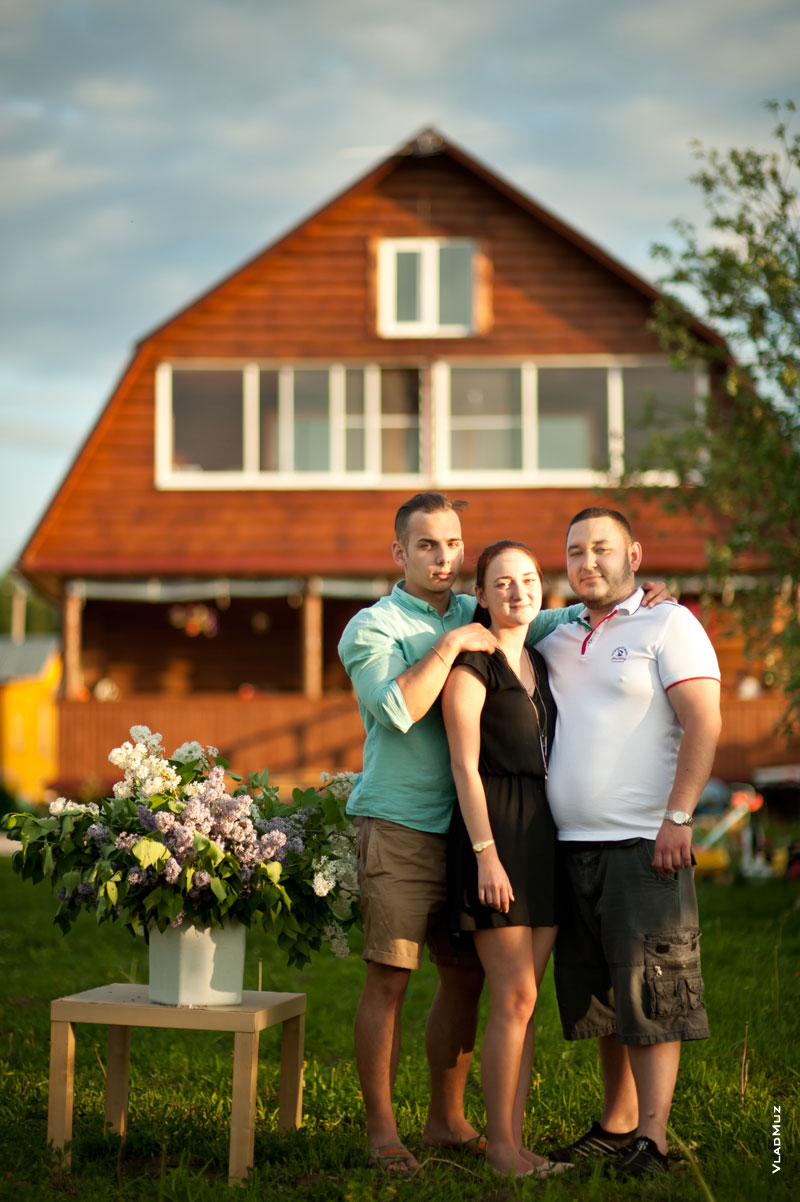Фото сестры и братьев в полный рост, рядом букет сирени на столике, вдали - деревянный дом