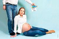 Фото беременной девушки сидя на студийном фоне, над ней будущий папа держит детский вертолет с надписью «Скоро буду»