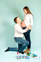 Студийное фото будущих родителей: мама стоит, папа перед ней на колене, на полу детские игрушки