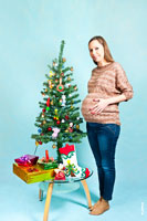 Фото беременной девушки стоя рядом с новогодней елкой в студии