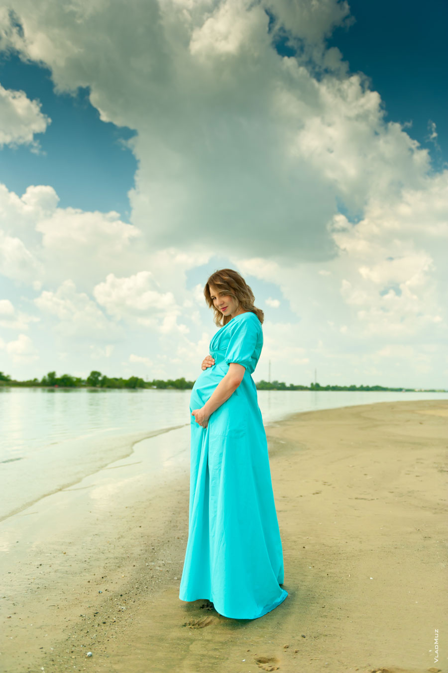 Фото беременной девушки в платье на песчаном берегу, на фоне высокого облачного неба