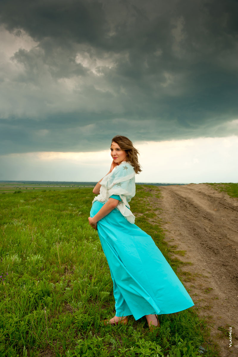 Фото беременной девушки в полный рост в бирюзовом платье на фоне грозового неба