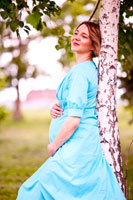 Фото беременной девушки в платье с руками на животе, прислонившись спиной к березе