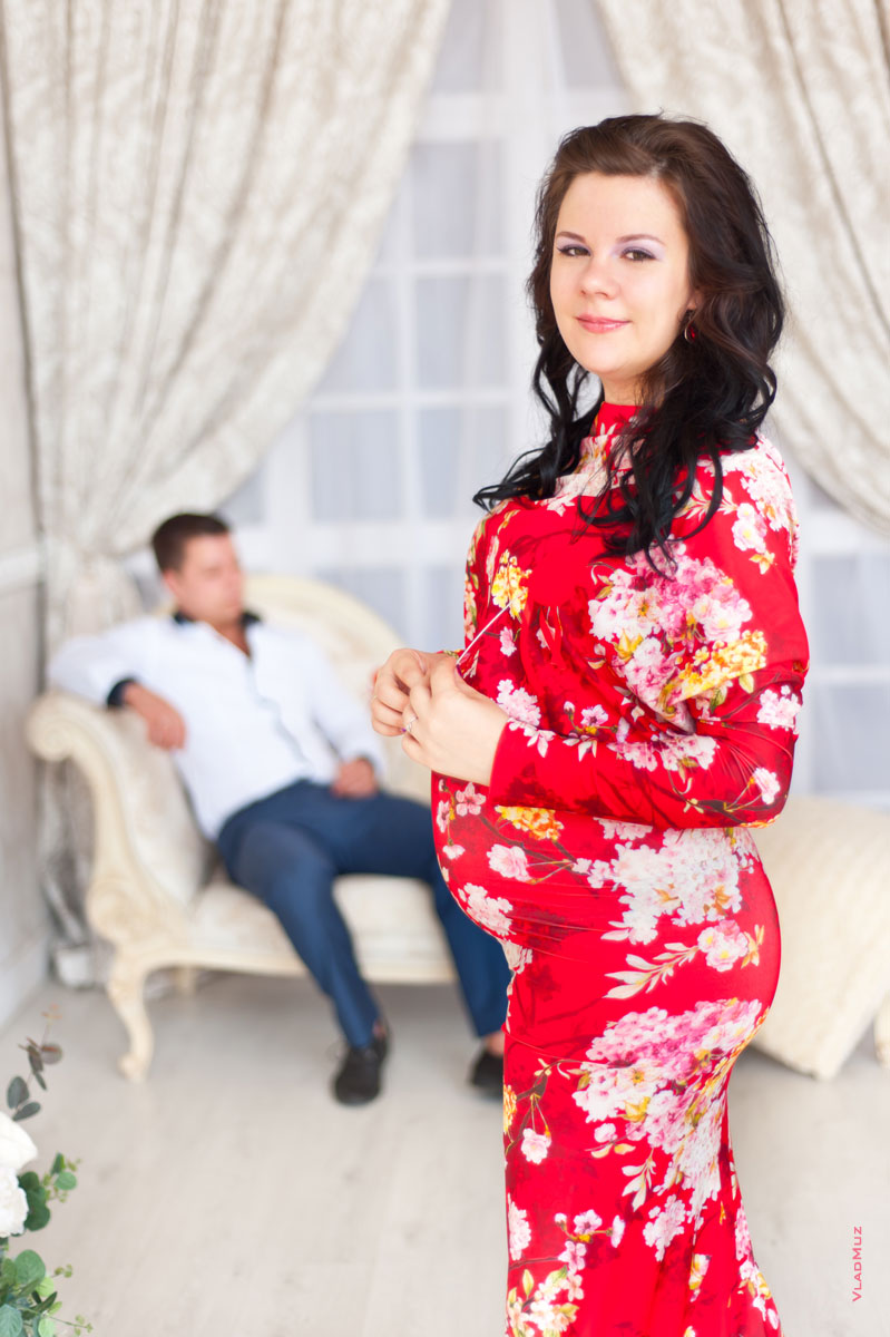 Фото беременной женщины в красном платье на переднем плане, вдали сидит мужчина в расфокусе