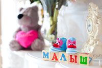 Фотонатюрморт: на деревянных кубиках набрано слово «Малыш», на кубиках стоят голубые пинетки, вдали — мягкая игрушка медведя с розовым сердцем
