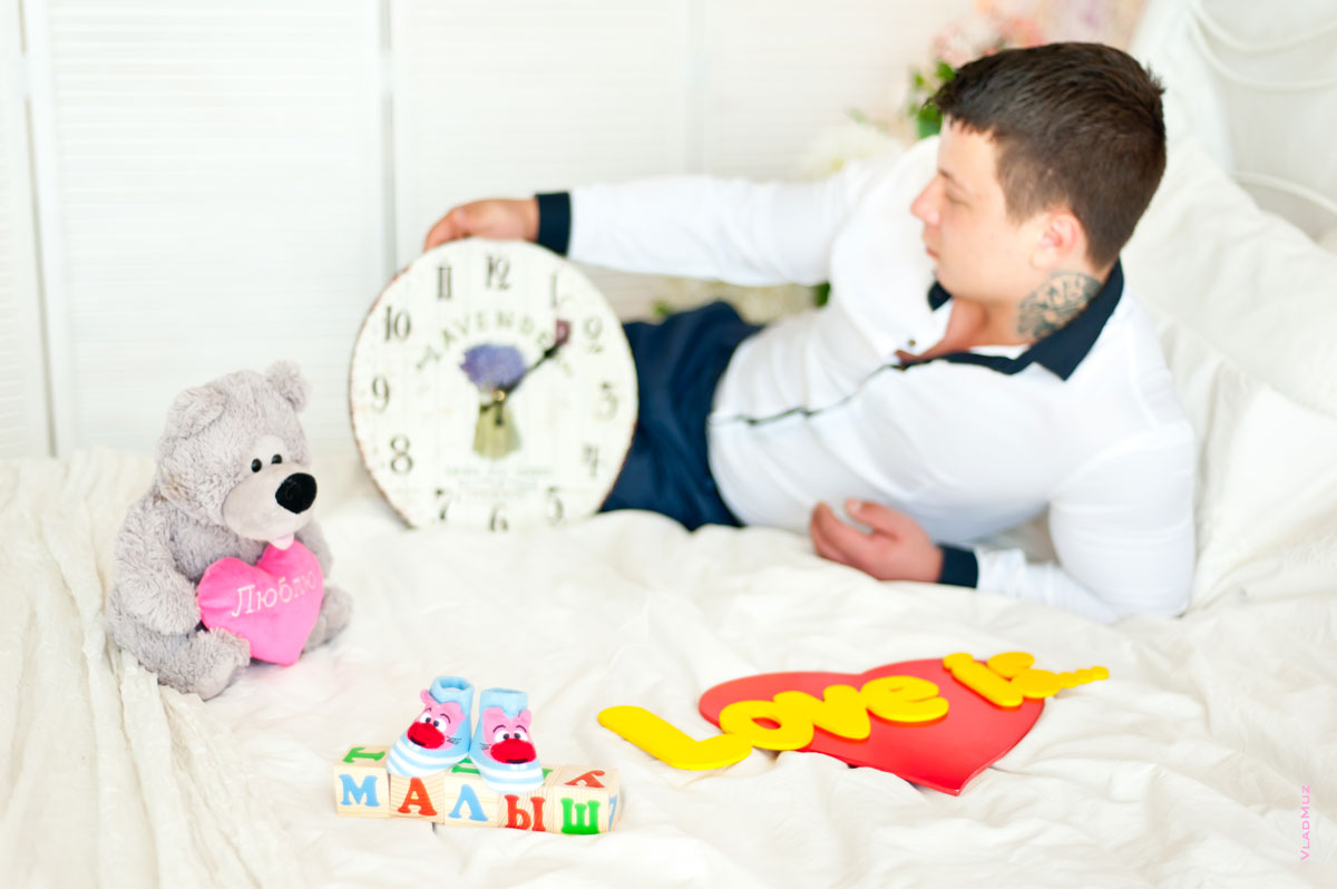 Фото будущего папы на кровати с часами в руках, рядом пинетки, слово «Малыш», мягкая игрушка и буквы “Love is”