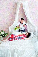 Семейное фото в спальне: на кровати лежит беременная жена в платье, перед ней стоит муж с цветами