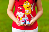 Фото беременной в красной яркой, веселой майке с голубыми пинетками в руках на фоне живота
