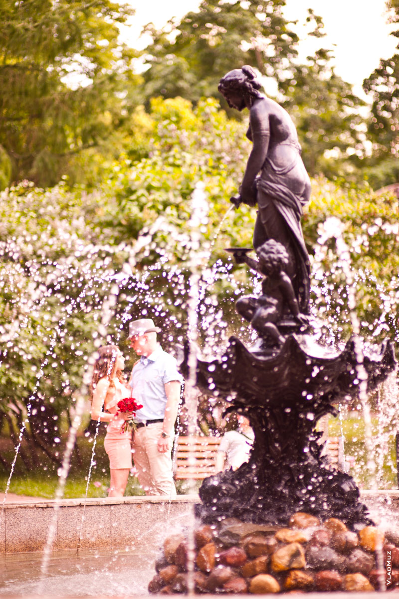 Здесь видно фонтан, черная фигура девушки льет вниз воду, сквозь струи и брызги воды вдали виднеется романтическая пара