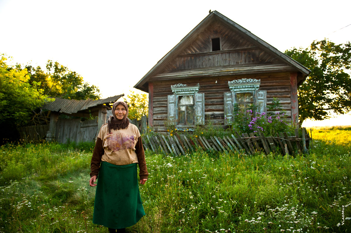 Жанровый фотопортрет сельской женщины в заброшенной деревне Белоруссии на фоне деревянного дома