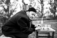 Черно-белый портрет мужчины в черной шапочке и в пальто с шарфом, на улице за столом, с бутылкой и стаканами