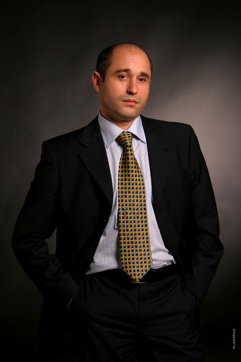 Студийный фотопортрет делового мужчины в костюме по пояс, на темном фоне, с руками в карманах