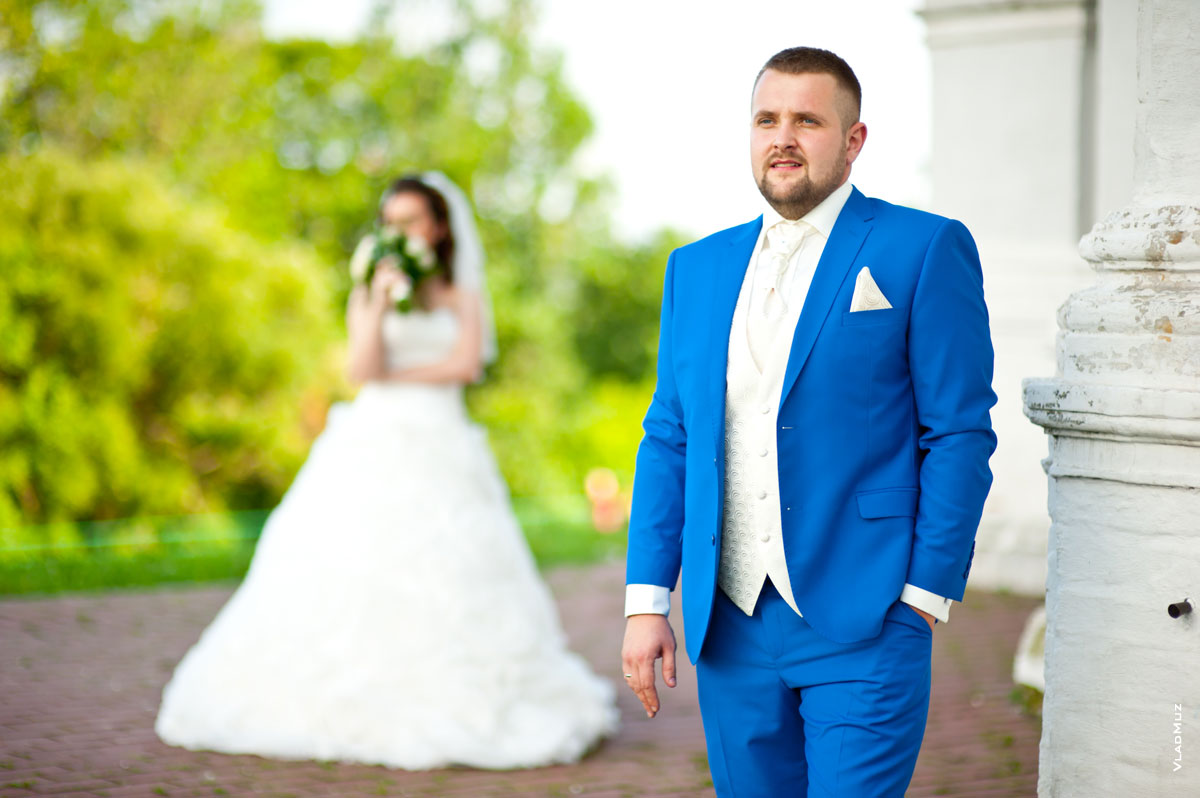 Свадебное фото жениха в фокусе, невеста с букетом стоит вдали в расфокусе