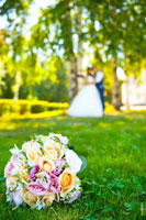 Фото свадебного букета в фокусе, лежащего на траве, на лужайке. Вдали, в расфокусе, стоят жених с невестой