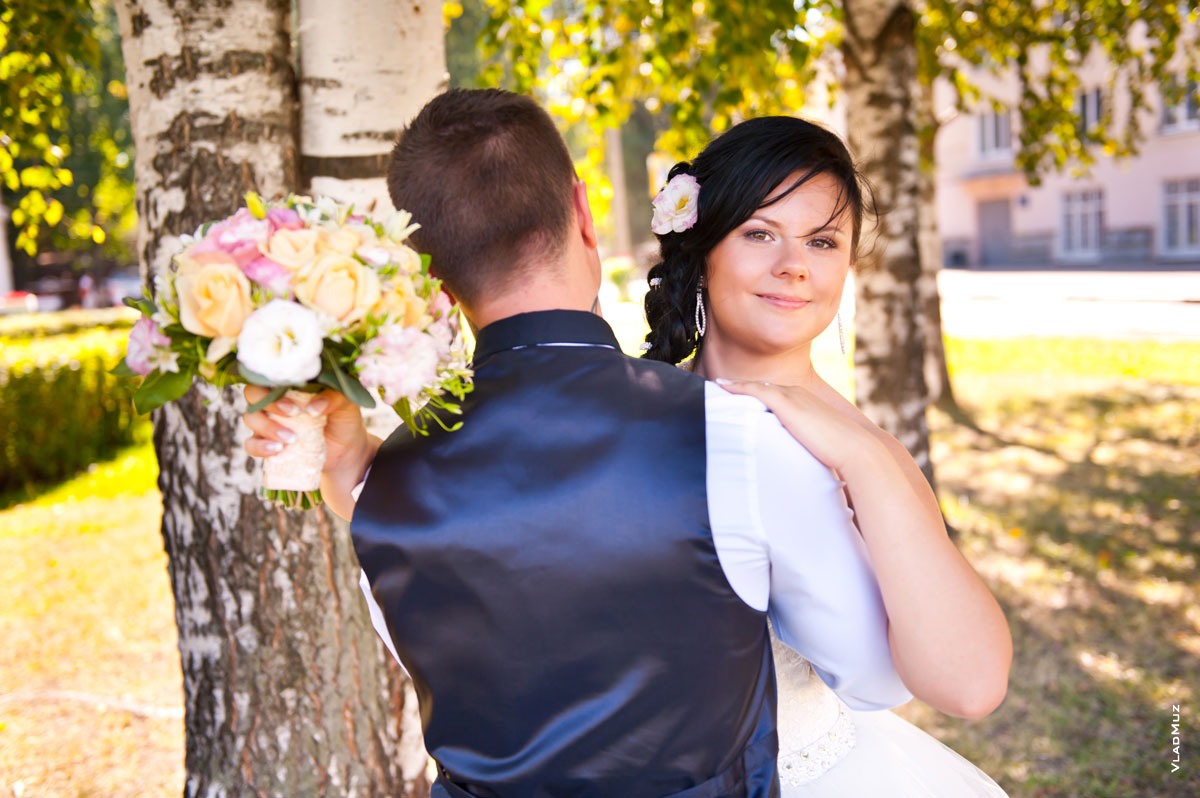 Игривое фото невесты с букетом в объятиях с женихом (жених стоит спиной, невеста смотрит в кадр)