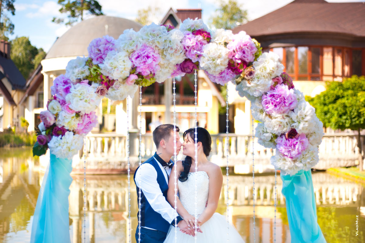 Фото жениха и невесты в свадебной арке из цветов, вдали в расфокусе — очертания отеля «Дворянское гнездо»