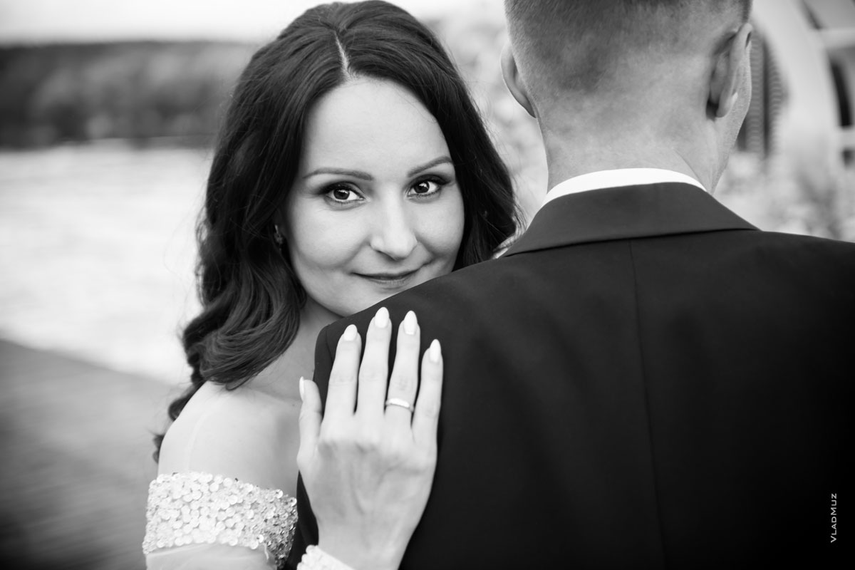 Ч/б фотопортрет невесты с акцентом на глазах и кольцом на руке, жених стоит спиной в кадре