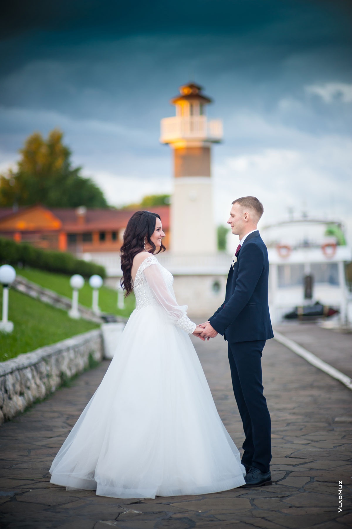 Фото жениха с невестой напротив друг друга, в полный рост, держащихся за руки, на фоне маяка комплекса «Белый берег»