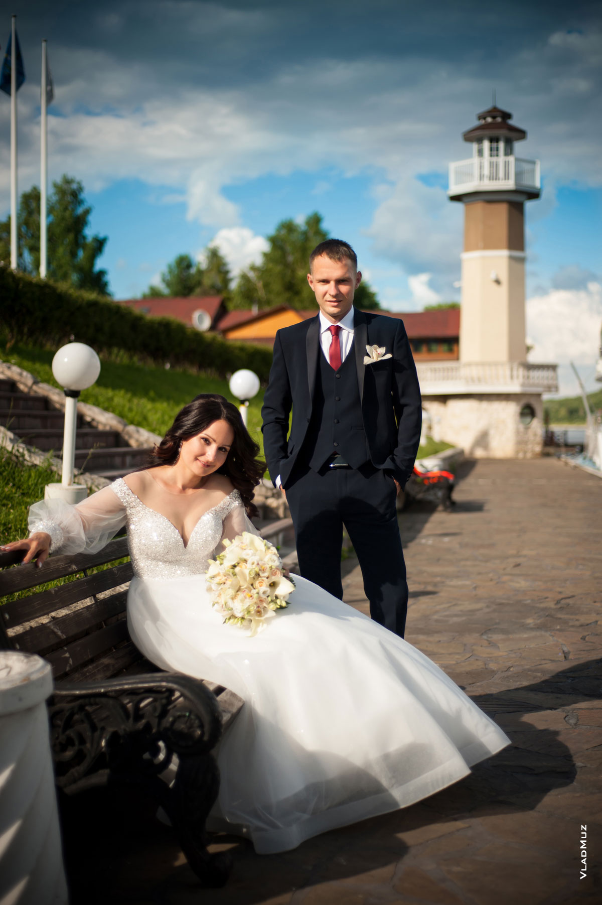 Фото невесты с букетом, сидя на лавочке, и жениха, стоя рядом (на фоне маяка ресторана «Белый берег»)