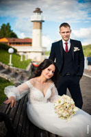Фото красивой и счастливой невесты, сидящей на лавочке, и жениха рядом, на фоне маяка ресторана «Белый берег»