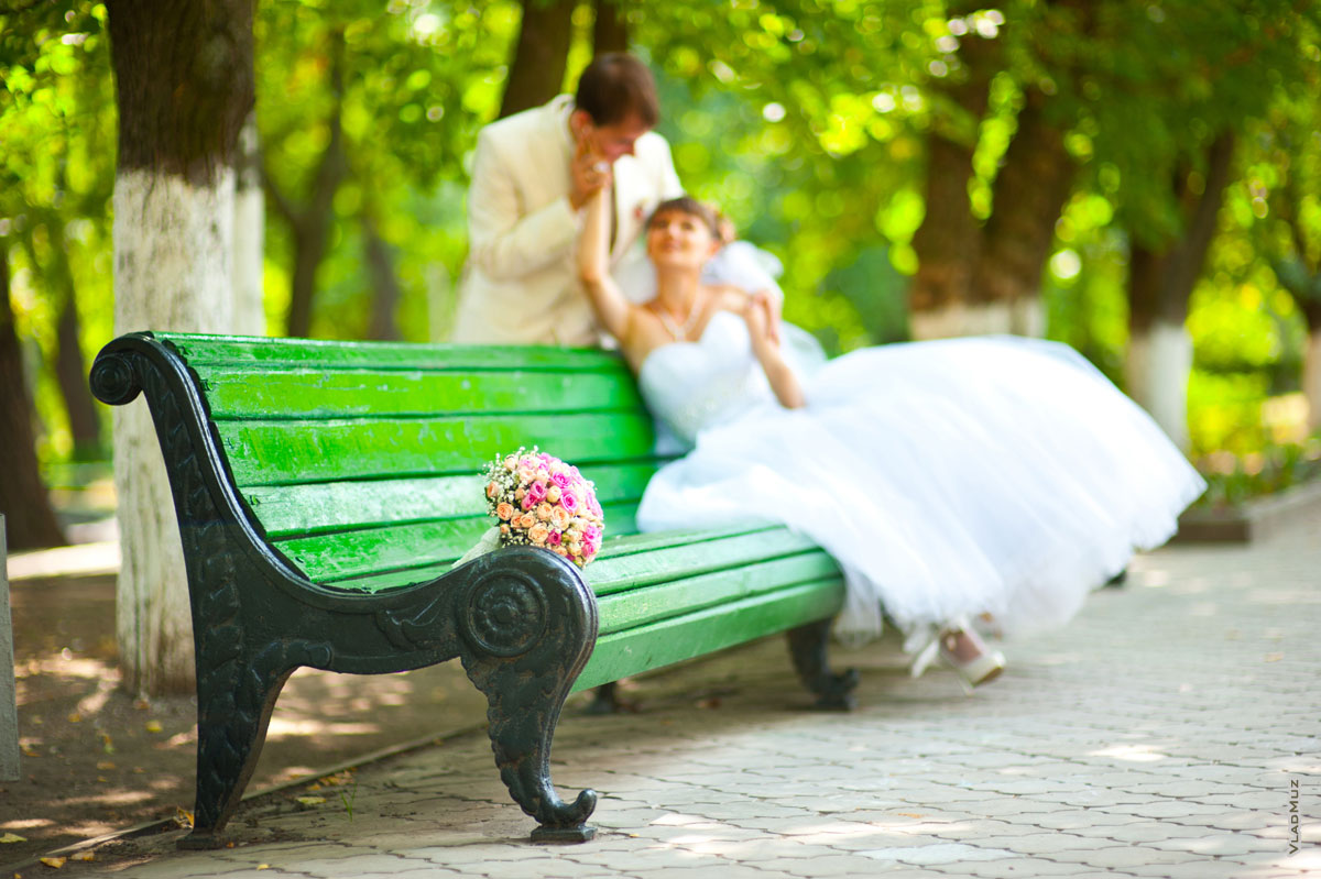Фото свадебного букета невесты на лавочке в фокусе, свадебная пара — в расфокусе