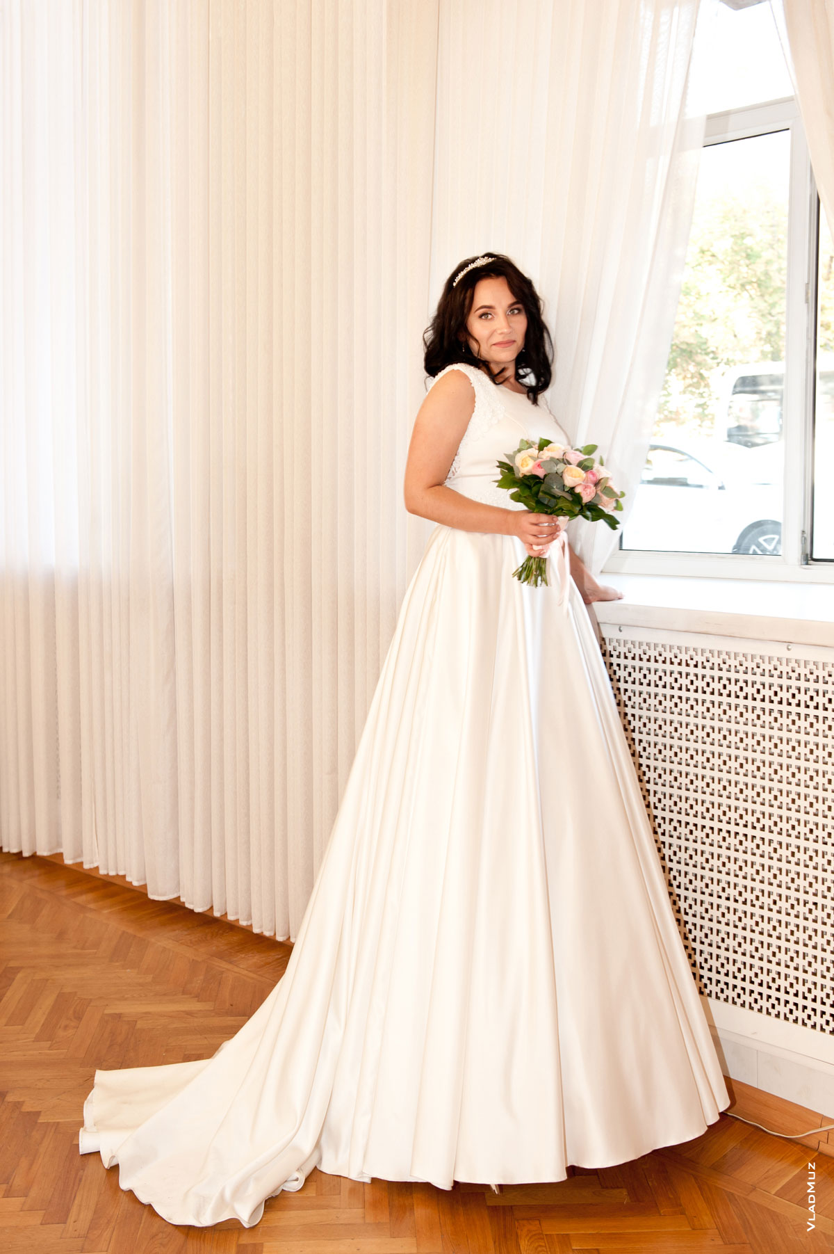 Фото невесты с букетом в длинном свадебном платье в полный рост, стоя у окна