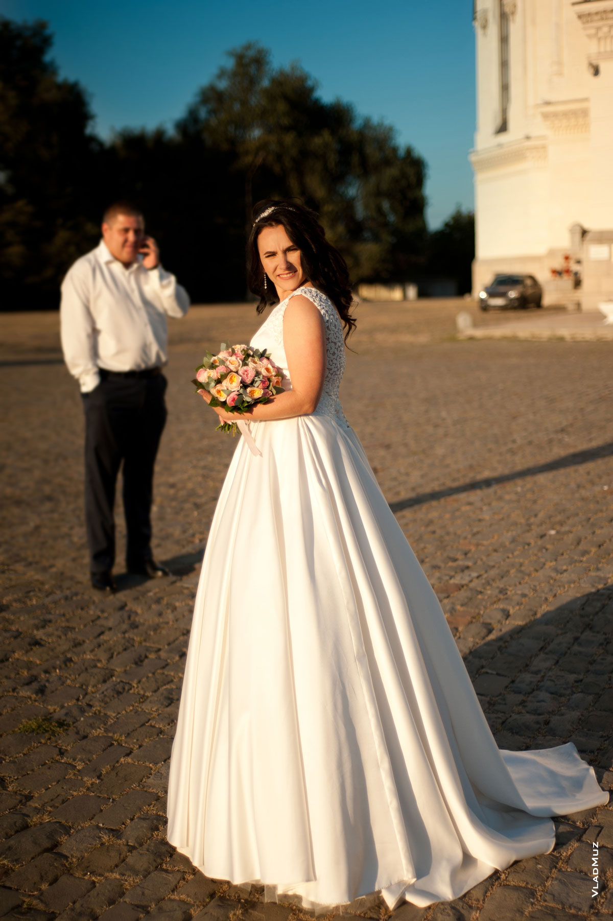 Фото невесты с букетом в фокусе, жених стоит вдали и говорит по телефону