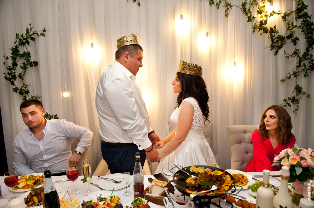 Фото жениха и невесты с коронами на головах и почетных свидетелей за свадебным столом в кафе